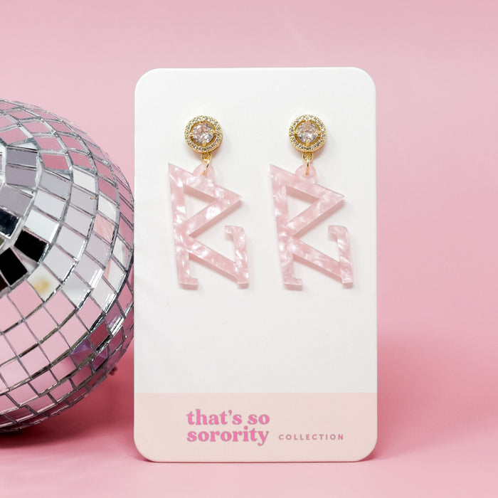 A pair of Delta Zeta Letter Pink Acrylic Earrings by The Darling Effect with gold and rhinestone studs are displayed on a white and pink card, perfect for bid day or sisterhood events—shown next to a silver disco ball on a pink background.