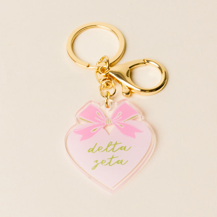 The Darling Effect’s Delta Zeta Sweetheart Bow Acrylic Keychain features a pastel bow, heart-shaped acrylic with green script, and a gold key ring and clasp—making it a charming sorority gift. Displayed on a light background.