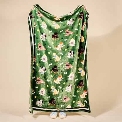 A person holds up The Darling Effect’s Dog Days Snuggle Up Plush Blanket, adorned with colorful cartoon dogs, bones, and balls; only their hands and slippered feet peek out from behind the cozy design.