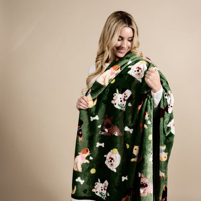 A smiling woman with long blonde hair wraps herself in The Darling Effect’s Dog Days Snuggle Up Plush Blanket, featuring playful cartoon dogs, bones, and tennis balls, against a plain beige background.