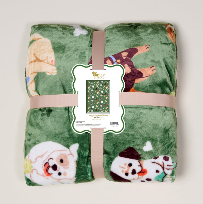 The Darling Effect’s Dog Days Snuggle Up Plush Blanket is green with playful cartoon dogs, rolled and tied with a beige ribbon. A front label shows the blanket pattern and product details.