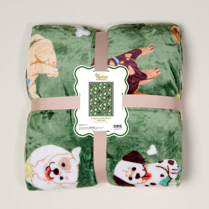 The Darling Effect’s Dog Days Snuggle Up Plush Blanket is green with playful cartoon dogs, rolled and tied with a beige ribbon. A front label shows the blanket pattern and product details.