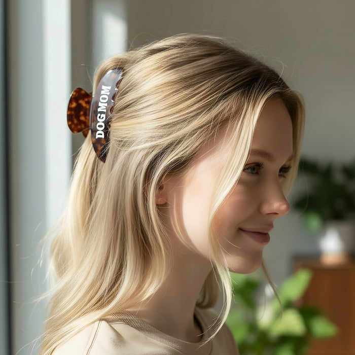 A smiling woman with long blonde hair wears The Darling Effect’s Dog Mom Word Up Claw Clip indoors. Soft light highlights her face, while green houseplants appear in the blurred background.