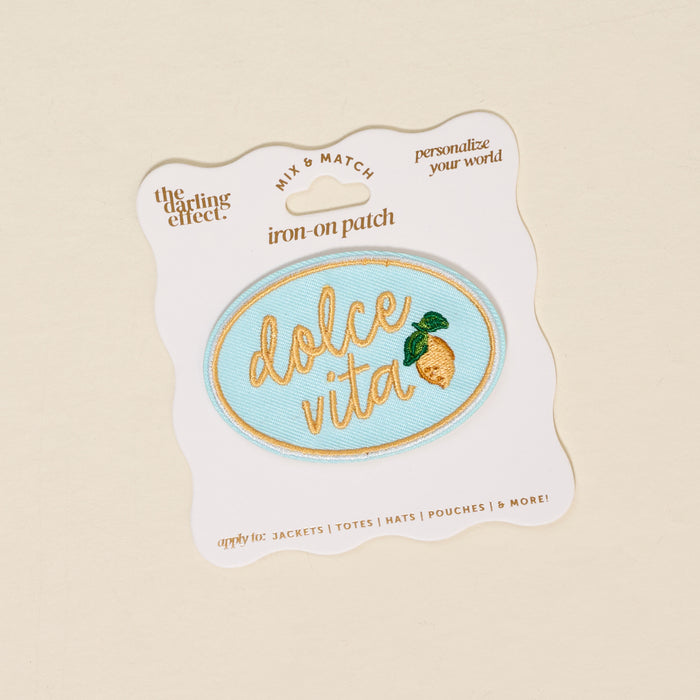 The Darling Effect Dolce Vita Mix & Match Iron-On Patch features an oval, light blue design with "dolce vita" in gold script and a lemon motif. Ideal for customizing jackets. Displayed on white backing with text.