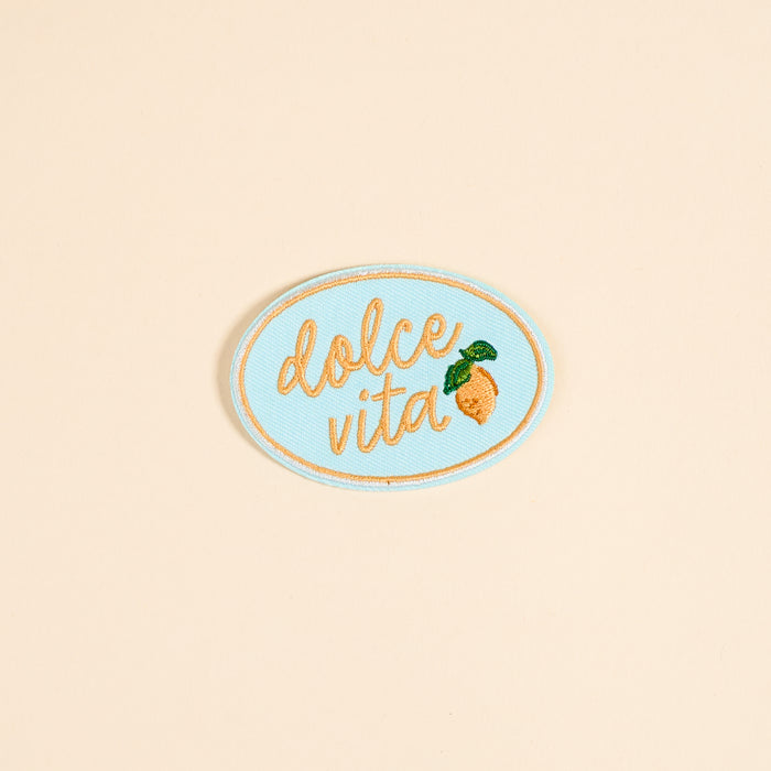 The Darling Effect's Dolce Vita Mix & Match Iron-On Patch features an oval shape, light blue background, gold cursive "dolce vita," and a yellow lemon with green leaves—perfect for brightening up your patch collection.