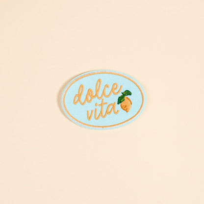 The Darling Effect's Dolce Vita Mix & Match Iron-On Patch features an oval shape, light blue background, gold cursive "dolce vita," and a yellow lemon with green leaves—perfect for brightening up your patch collection.