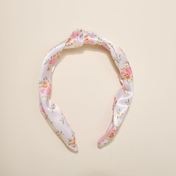 The Darling Effect’s Endless Daydream Cream Classic Twist Headband features a white satin finish with pink, orange, and green floral patterns—an elegant, versatile accessory for any occasion, shown on a beige background.
