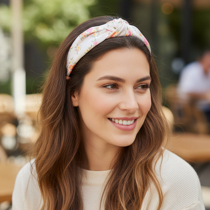 A woman with long brown hair smiles while sitting outdoors at a café, wearing The Darling Effect's Endless Daydream Cream Classic Twist Headband and a cream-colored sweater.