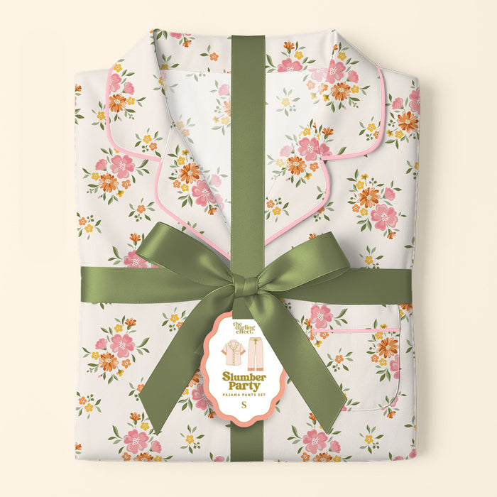 A folded set of Endless Daydream Cream Slumber Party Pajama Pants by The Darling Effect, featuring a pink and orange floral print on ultra-soft fabric, tied with a green ribbon and gift tag. Gift-ready for any occasion.