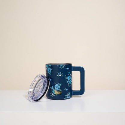 The Darling Effect’s Endless Daydream Navy 14 oz Insulated Mug with a blue floral pattern and square handle sits on a white surface, its transparent lid propped against it. The background is plain and light-colored.