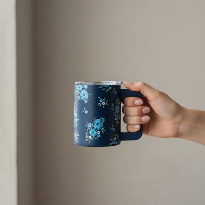 A manicured hand holds The Darling Effect Endless Daydream Navy 14 oz Insulated Mug, featuring a pattern of light blue and white flowers, against a plain, light background.