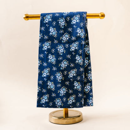 The Endless Daydream Navy Tea Towel by the-darling-effect, featuring small light blue flowers and green leaves, is draped neatly over a gold metal stand against a plain light background.
