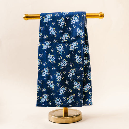 The Endless Daydream Navy Tea Towel by the-darling-effect, featuring small light blue flowers and green leaves, is draped neatly over a gold metal stand against a plain light background.