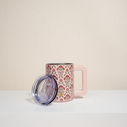 The Darling Effect’s Everbloom Dusty Rose 14 oz Insulated Mug, featuring a floral pattern and large handle, sits on a white surface with its clear plastic lid beside it. The plain light background highlights this charming drinkware.