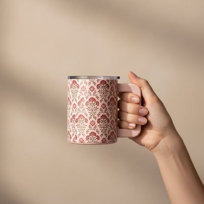 A hand with manicured nails holds The Darling Effect’s Everbloom Dusty Rose 14 oz Insulated Mug, featuring a red and gray floral design, set against a neutral beige background with soft shadows.
