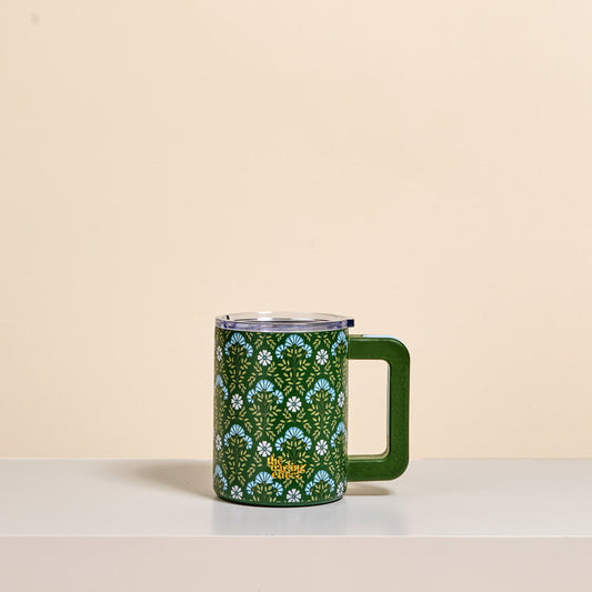The Darling Effect's Everbloom Forest Green 14 oz Insulated Mug features double-wall insulation and a floral pattern of blue and white flowers, displayed on a white shelf with a beige background.