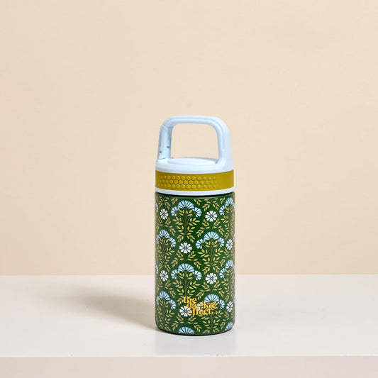 The Darling Effect’s Everbloom Forest Green 6 oz Lil' Bitty Bottle features a light blue handle and lid, a green body with white and yellow florals, and is shown on a white surface against a beige background.