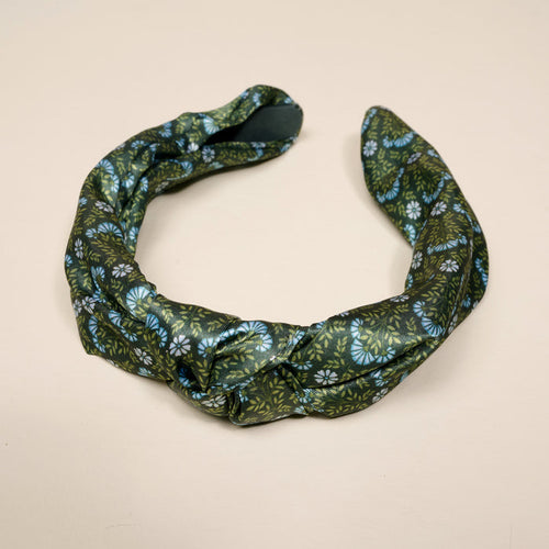 The Darling Effect's Everbloom Forest Green Classic Twist Headband features a dark green base with blue and light green florals, an elegant knot detail, and is shown on a light beige background.