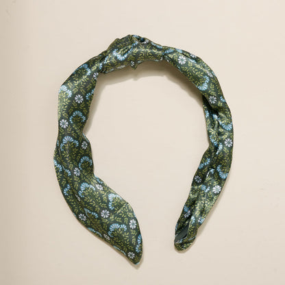 The Darling Effect Everbloom Forest Green Classic Twist Headband, featuring a white and blue floral pattern and a stylish knot detail, is displayed against a plain beige background.