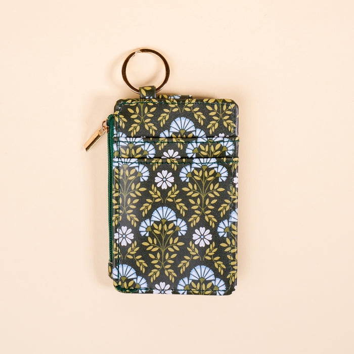 The Darling Effect’s Everbloom Forest Green Keychain Wallet features a white and light blue floral pattern, gold zipper, and keyring—a slim, functional wallet for floral lovers—shown on a neutral beige background.