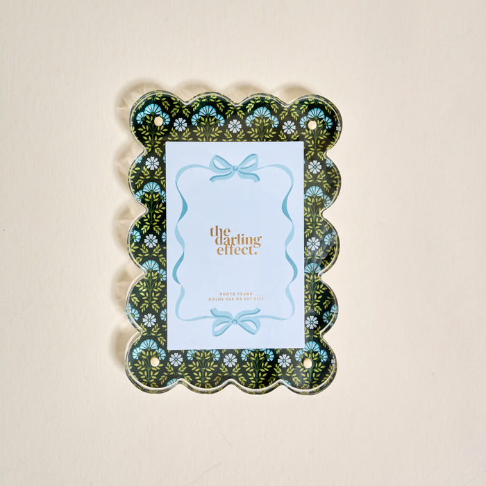 The Everbloom Forest Green Rectangle Scalloped Acrylic Picture Frame by The Darling Effect displays a card with a decorative blue bow, features a black background with light blue floral accents, and sits on a beige surface.