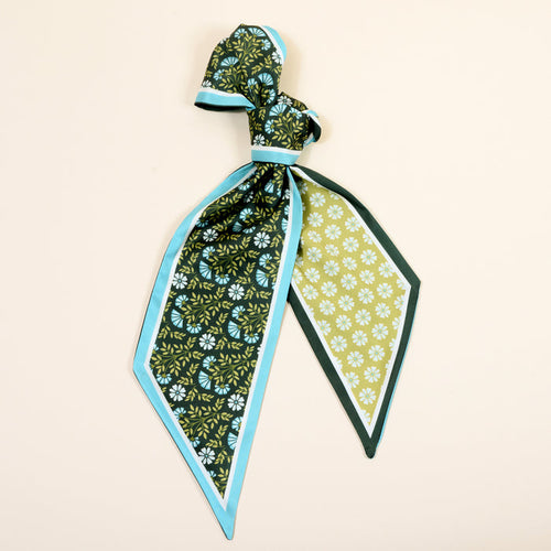 The Darling Effect's Everbloom Forest Green Twirl & Tie Scarf Accessory features green, yellow, and white florals with blue borders. Styled in a loose knot on beige—an endlessly chic and versatile accent for any look.