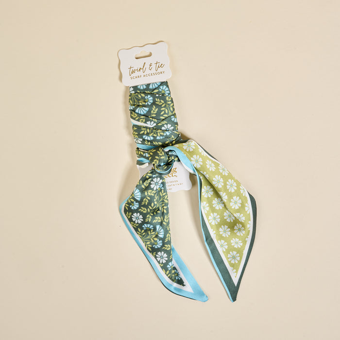 The Darling Effect’s Everbloom Forest Green Twirl & Tie Scarf Accessory features white floral patterns and a light blue border, neatly folded on a branded card—a versatile piece for endless styling options.