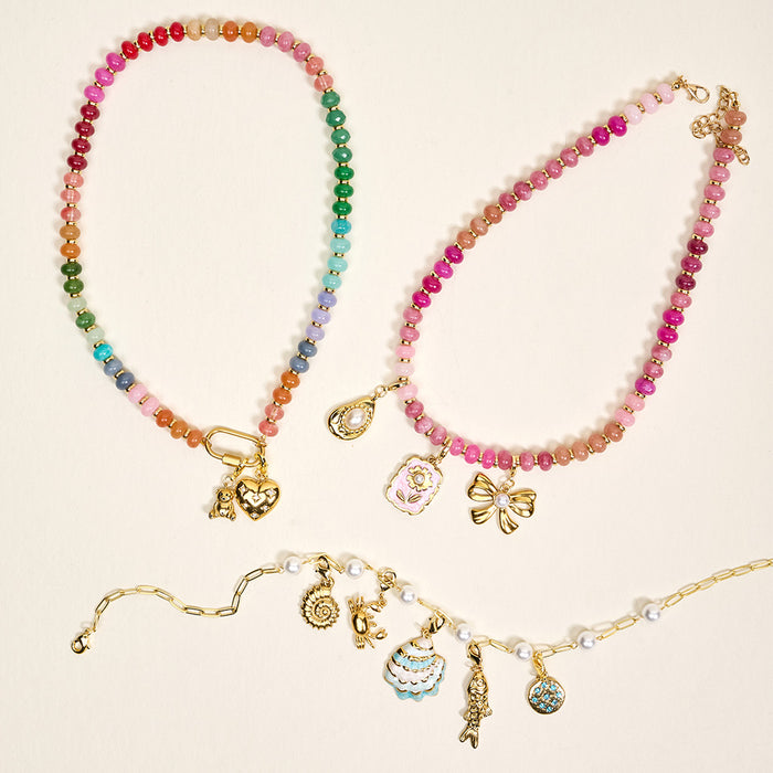 Three colorful necklaces by The Darling Effect: a rainbow beaded one with heart and teddy bear charms, a pink beaded necklace with a seashell and bow, and a gold chain featuring the Fish Jewelry Charm Gold, ideal for layering.