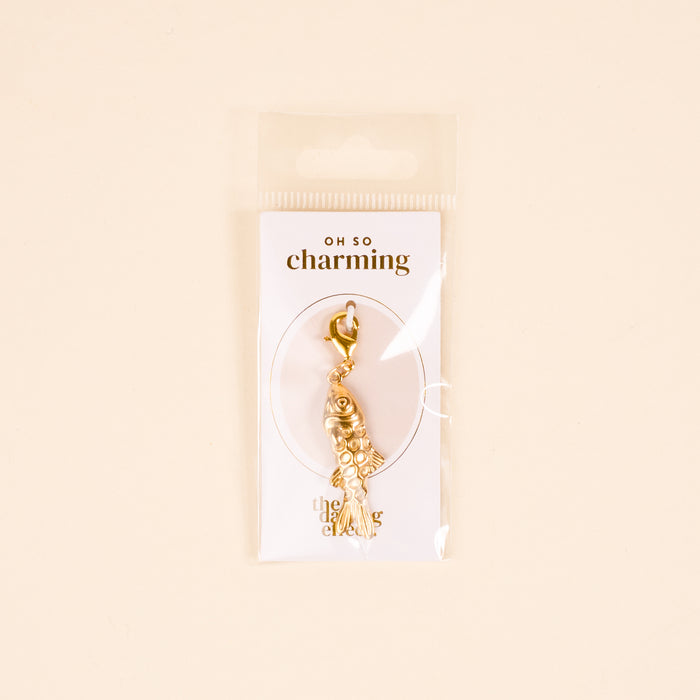 The Darling Effect Fish Jewelry Charm Gold features intricate detailing, comes attached to an "Oh So Charming" card, and is packaged in a clear sleeve against a light beige background—perfect for your charm bar.