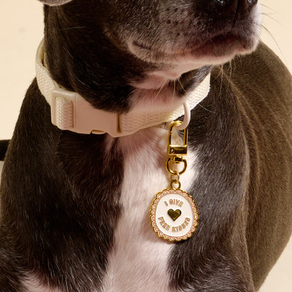 A close-up of a dog wearing The Darling Effect's cream collar with the Free Kisses Pawfect Collar Charm, which is round, gold and white, reads "GIVE FREE KISSES" with a heart detail. The dog's face is partly out of frame, showcasing this cute pet accessory.
