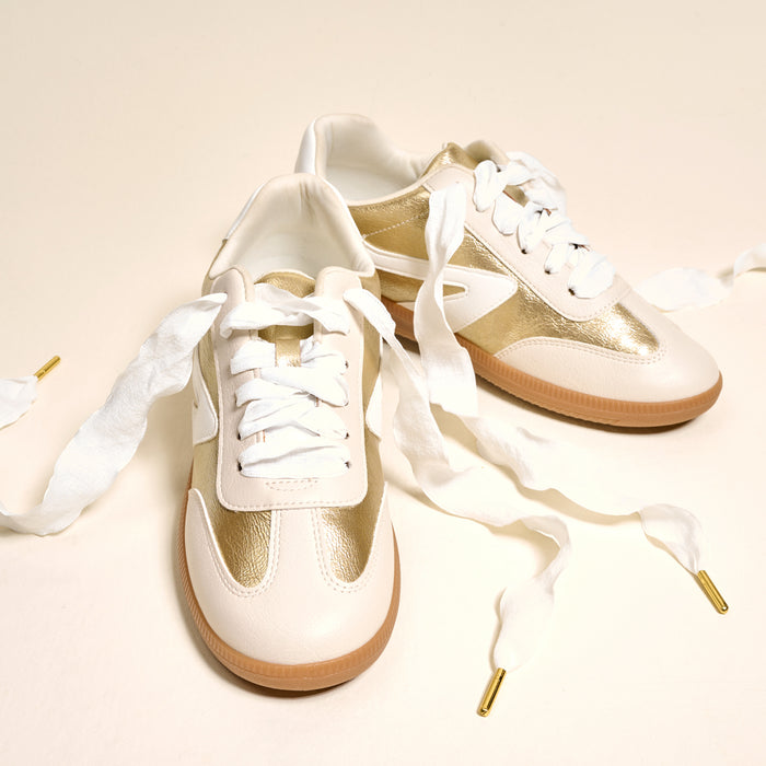 A pair of cream and gold sneakers with durable gold-tipped laces is displayed on a cream background, adorned with elegant Frilly White Step It Up Shoelaces by The Darling Effect.