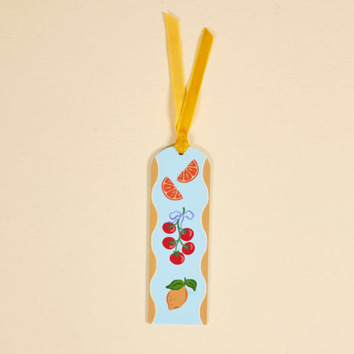 The Fruit Market Acrylic Bookmark features a rectangular shape with a yellow ribbon, showcasing illustrations of orange slices, tomatoes on the vine with a blue bow, and a mango on a light blue background with a wavy beige border—perfect for book lovers.