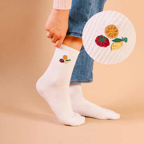 A person wears The Darling Effect’s Fruit Market Embroidered Crew Socks—white ribbed socks with playful orange, strawberry, and pineapple designs—standing on a beige surface, with a close-up circle showing the vibrant embroidered fruit detail.