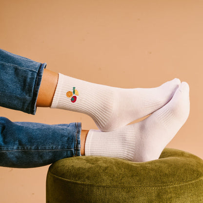 A person wearing The Darling Effect Fruit Market Embroidered Crew Socks, featuring colorful fruit designs, relaxes on a green ottoman. They are in blue jeans with a beige background.