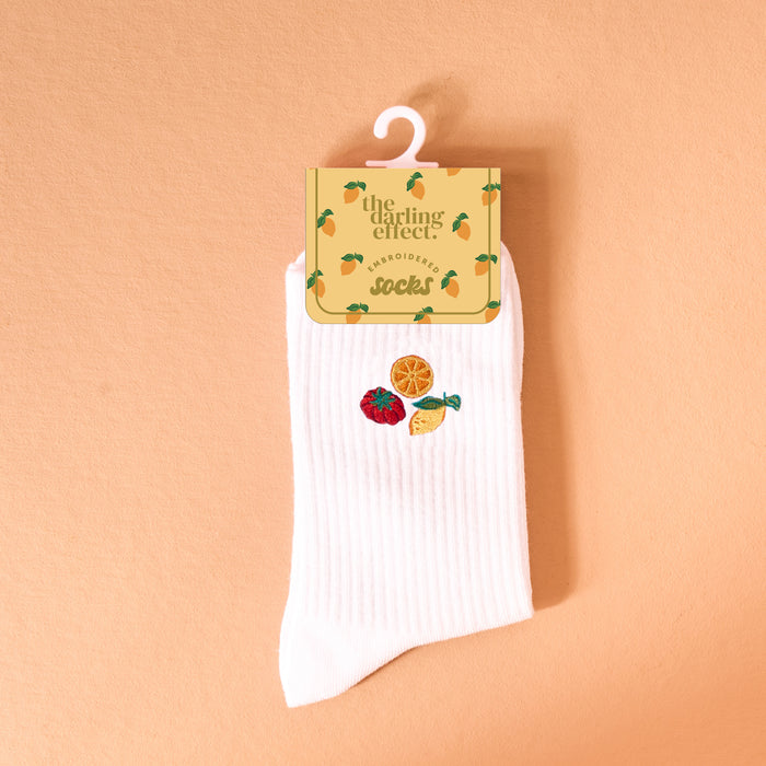 The Darling Effect Fruit Market Embroidered Crew Socks: white women’s socks with a ribbed texture, featuring colorful embroidered strawberry, orange slice, and lemon wedge designs, shown against a peach background.
