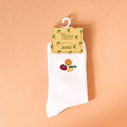 The Darling Effect Fruit Market Embroidered Crew Socks: white women’s socks with a ribbed texture, featuring colorful embroidered strawberry, orange slice, and lemon wedge designs, shown against a peach background.