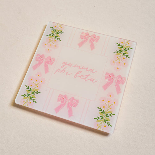 The Darling Effect's Gamma Phi Beta Acrylic Coaster features pink script, pink bows, and yellow flowers with green leaves on a light background—an adorable sorority gift.