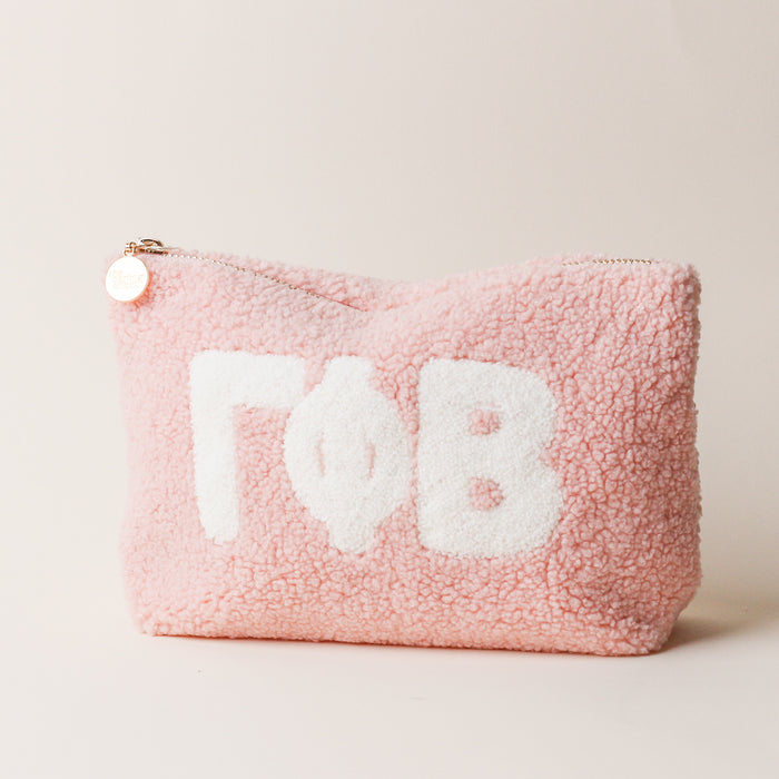 The Darling Effect Gamma Phi Beta Blush Teddy Letter Pouch is a pink sherpa pouch with large white Greek letters, a zipper topped with a gold charm, and is perfect as a cute carry-on accessory. The background is off-white.
