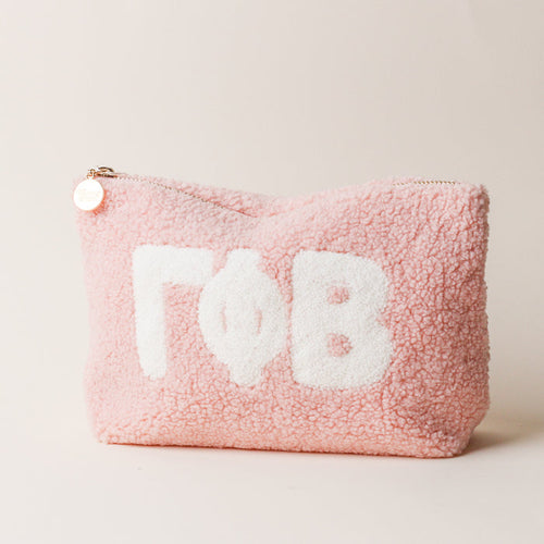 The Darling Effect Gamma Phi Beta Blush Teddy Letter Pouch is a pink sherpa pouch with large white Greek letters, a zipper topped with a gold charm, and is perfect as a cute carry-on accessory. The background is off-white.