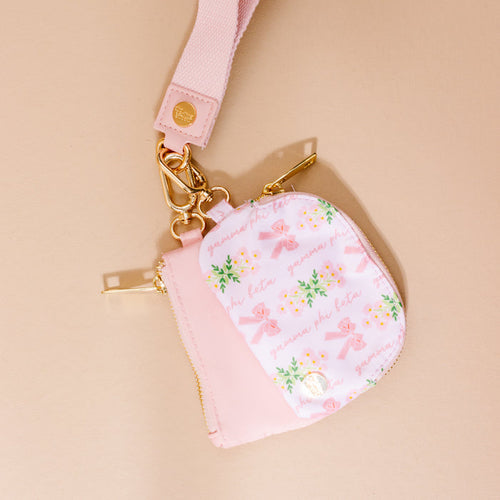 The Darling Effect’s Gamma Phi Beta Dynamic Duo Pouch Wristlet in pink and white features floral and ribbon patterns, gold hardware, a detachable strap, and “gamma phi beta” text. Displayed on a beige surface.
