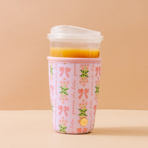 The Darling Effect's Gamma Phi Beta Large Sip Savvy Drink Sleeve, featuring floral patterns, bows, and cursive on beige, wraps a plastic cup with orange juice and a clear lid—perfect for sorority gifts.