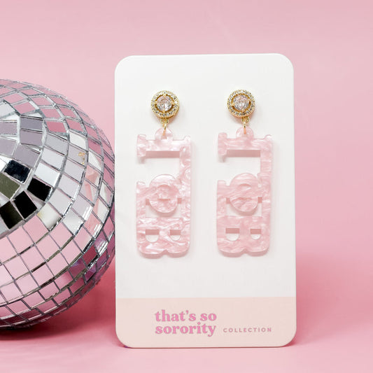 A pair of Gamma Phi Beta Letter Pink Acrylic Earrings by The Darling Effect, featuring gold and rhinestone studs, displayed on a "that's so sorority collection" card beside a silver disco ball—perfect for sisterhood events or bid-day baskets.