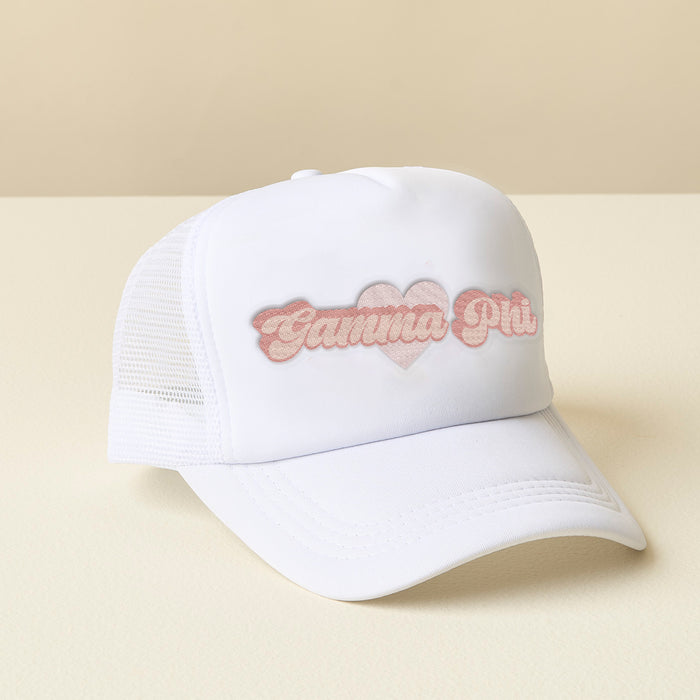 The Darling Effect Gamma Phi Beta White Trucker Hat features a pink heart patch with "Gamma Phi" in pink cursive. Stylish for sorority gifts, it’s shown on a light surface against a beige background.