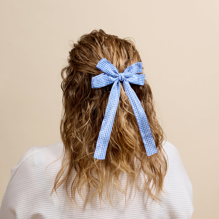 A person with wavy, shoulder-length hair styled half-up and tied with The Darling Effect’s Gingham Blue Double Take Hair Bow, shown from the back against a plain beige background—a charming statement accessory.