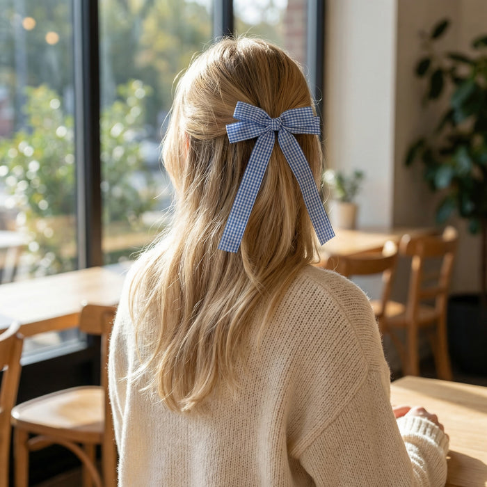 A woman with long blonde hair sits at a sunlit wooden table, facing away as she wears The Darling Effect's Gingham Blue Double Take Hair Bow with a cream sweater, showcasing her charming accessory indoors.