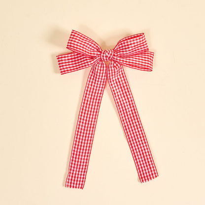 The Darling Effect's Gingham Red Double Take Hair Bow features a red and white checkered ribbon tied into a bow with two long tails, perfect for hair accessories or clips, shown on a plain beige background.