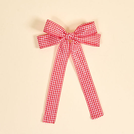 The Darling Effect's Gingham Red Double Take Hair Bow features a red and white checkered ribbon tied into a bow with two long tails, perfect for hair accessories or clips, shown on a plain beige background.