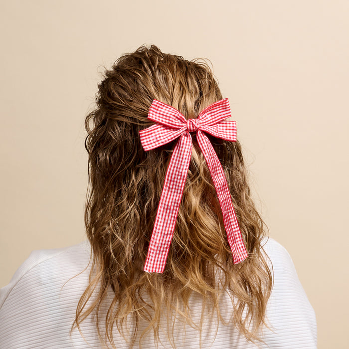 A person with wavy, light brown hair styled half-up wears The Darling Effect’s Gingham Red Double Take Hair Bow, shown from behind against a plain beige background.