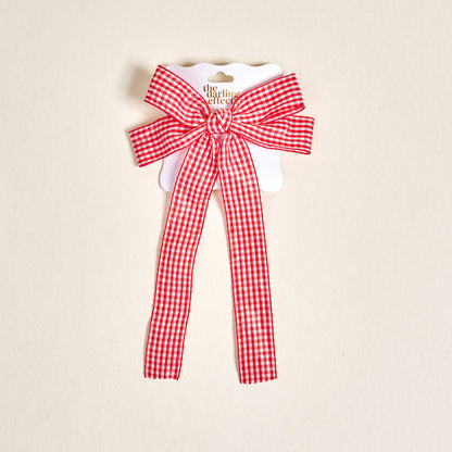 The Gingham Red Double Take Hair Bow by The Darling Effect features a red and white checkered fabric with two long tails, perfect for hair accessories, displayed on a white card against a light beige background.