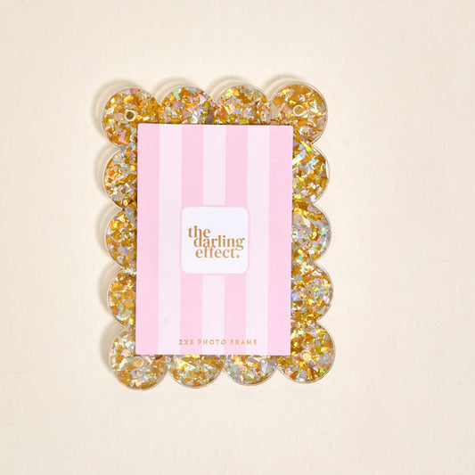 The Darling Effect Gold Glitter Mini Acrylic Picture Frame Magnet features a rectangular design with round, gold glitter-filled discs as the border. It displays a pink and white striped card on a plain, light background.
